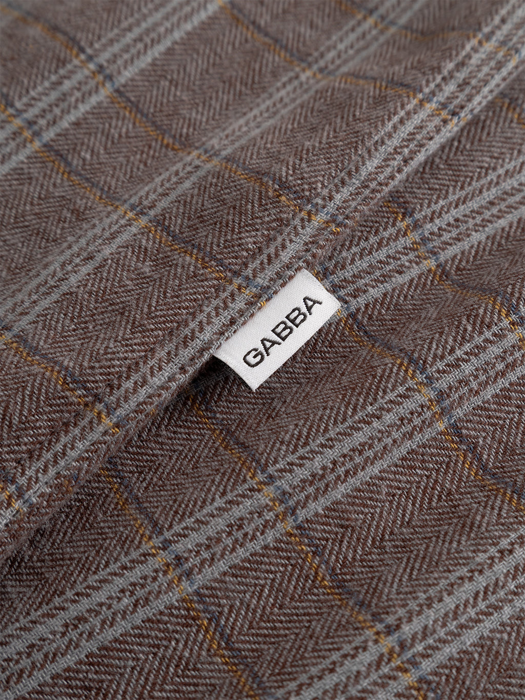Close-up of the Sven Reg Herri LS - Brown Rust shirt fabric in brown rust and gray plaid herringbone, with a small white Gabba label sewn into the seam.