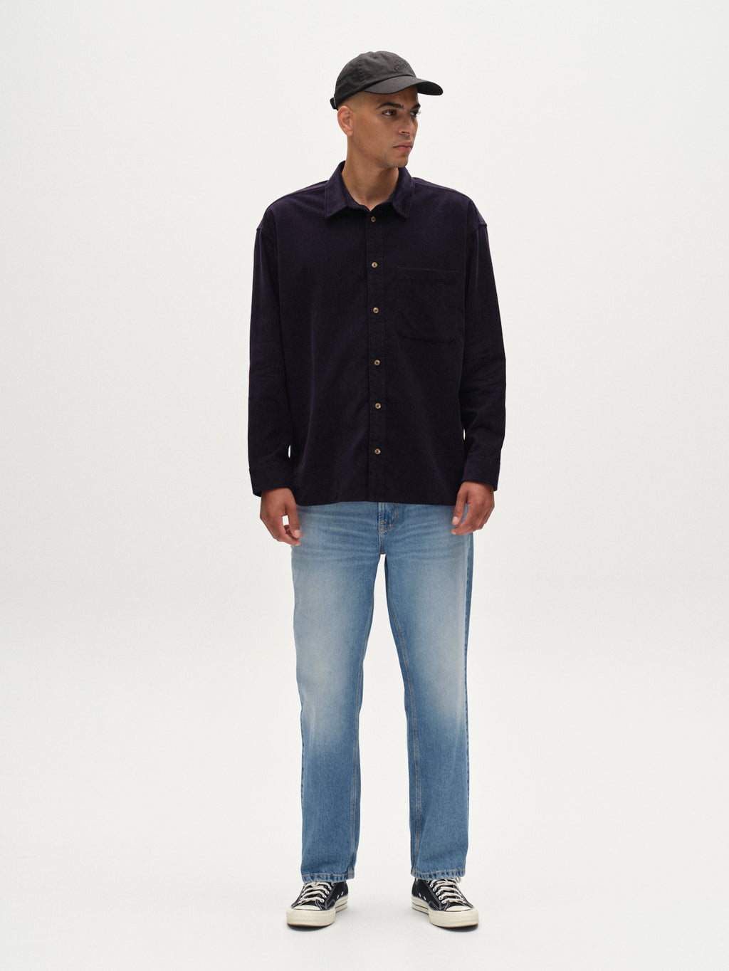 A man wearing the Gabba Jens Over Cord LS - Night Sky overshirt, a black cap, light blue jeans, and black-and-white sneakers stands facing forward against a plain white background.
