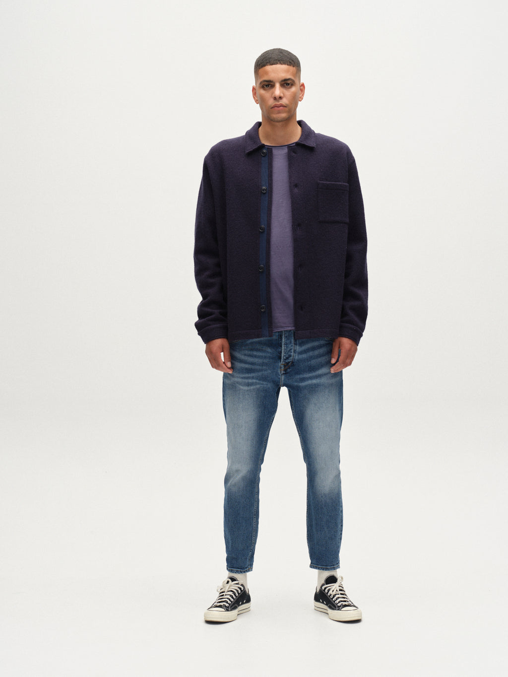 A man stands facing forward against a plain white background, wearing Gabbas Alex Cros - Mid Blue Denim jeans, a dark button-up jacket over a t-shirt, and black high-top sneakers. His arms are relaxed at his sides.