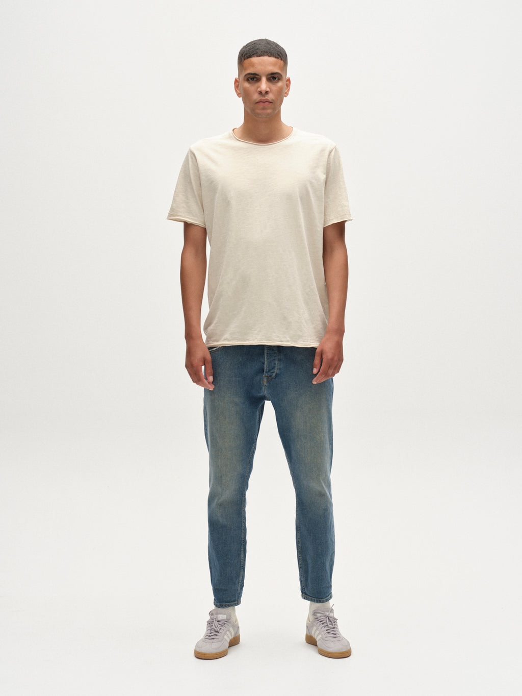 A man faces forward against a plain white background, wearing a beige short-sleeve t-shirt, Gabba Alex Ulio Mid Blue Denim relaxed tapered jeans for stretch comfort, and light-colored sneakers. His arms rest naturally at his sides.