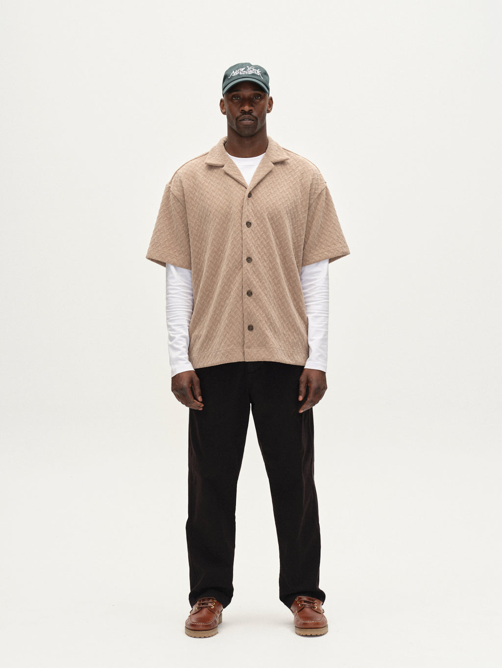 A man faces forward against a plain white background, wearing the Gabba Jens Over Diam SS overshirt in Sand over a white long-sleeve shirt, with black pants, brown shoes, and a green cap.