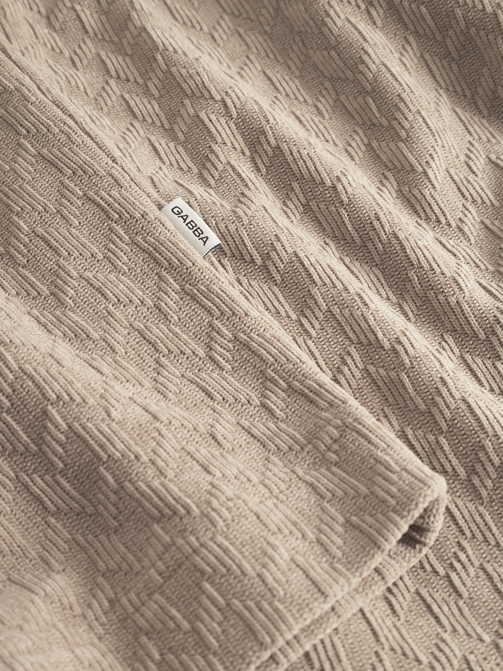 A close-up of the textured Sand Jens Over Diam SS overshirt by Gabba, showing its subtle geometric pattern and a small white Gabba tag sewn onto the beige fabric.