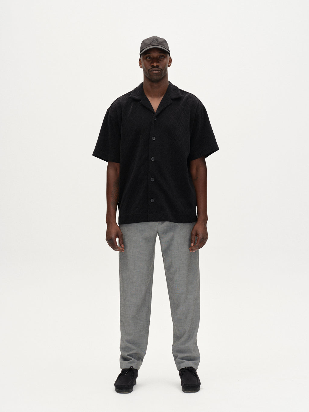 A man faces forward against a plain white background, wearing the Gabba Jens Over Diam SS shirt in black, light gray pants, black shoes, and a dark baseball cap.
