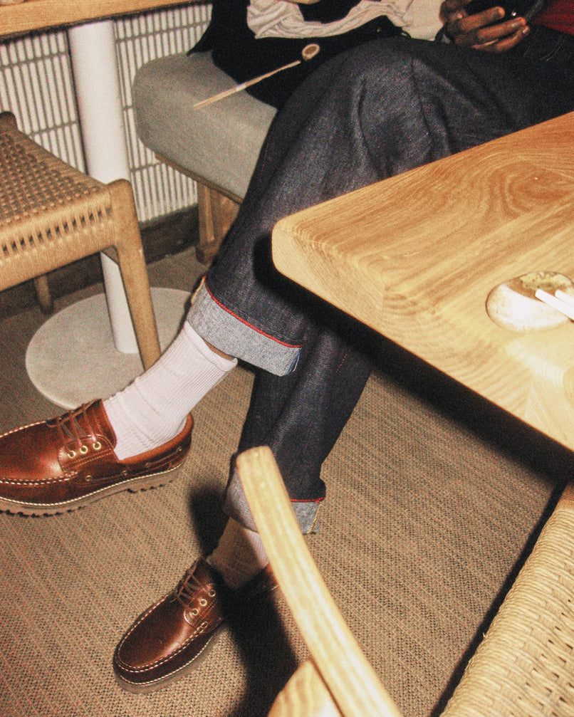 Someone wearing Gabba’s David Chev - Dark Navy jeans with cuffs, white socks, and brown leather shoes sits at a wooden table. Woven chairs and a herringbone beige carpet are visible; theyre holding a phone, upper body partly out of frame.