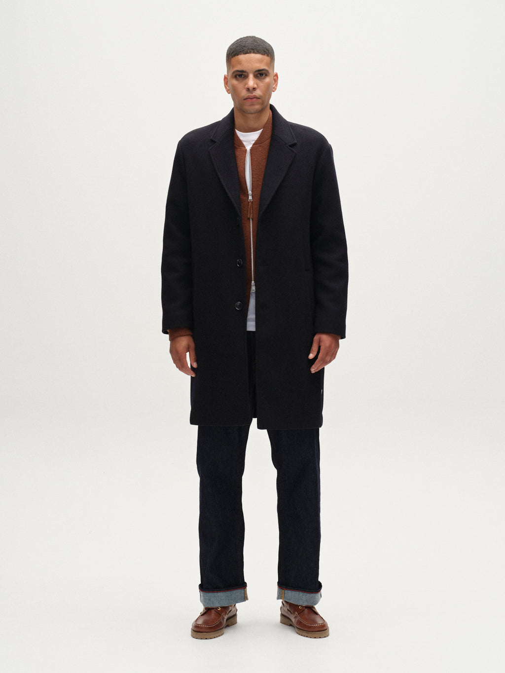 A man stands facing forward, wearing Gabba's Leeds Drift - Navy oversized wool coat over a brown sweater, white shirt, dark jeans, and brown shoes against a plain white background.