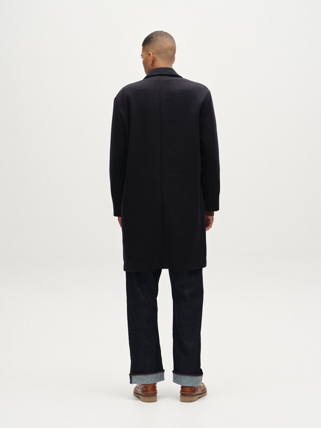 A person faces away from the camera, wearing Gabba's Leeds Drift - Navy oversized wool coat, dark pants, and brown shoes against a plain white background.