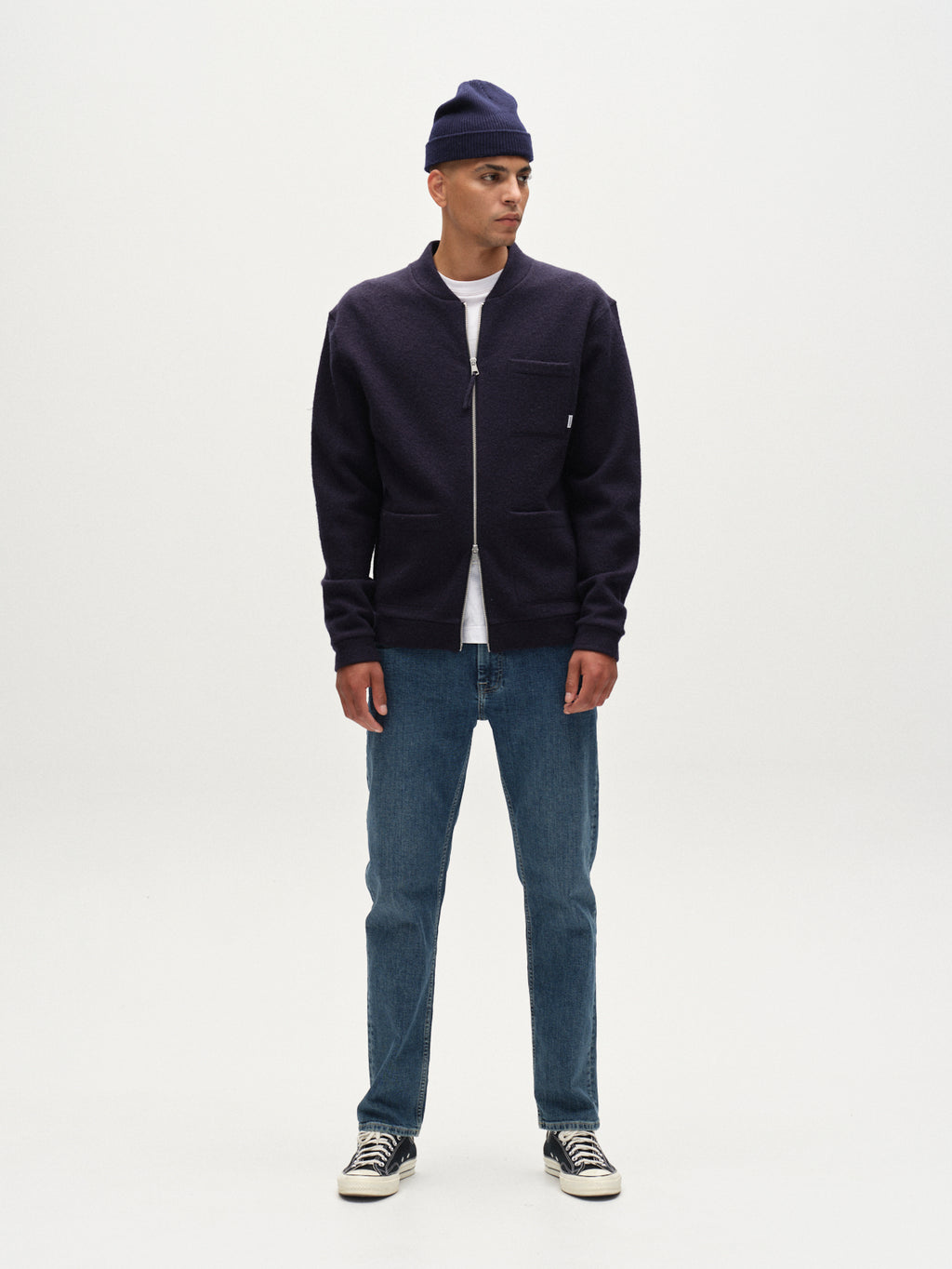 A man stands facing forward, wearing a navy beanie, navy zip-up jacket, white T-shirt, Gabba Marc Jingo dark blue denim jeans (comfort stretch straight-leg), and black-and-white sneakers against a plain white background.