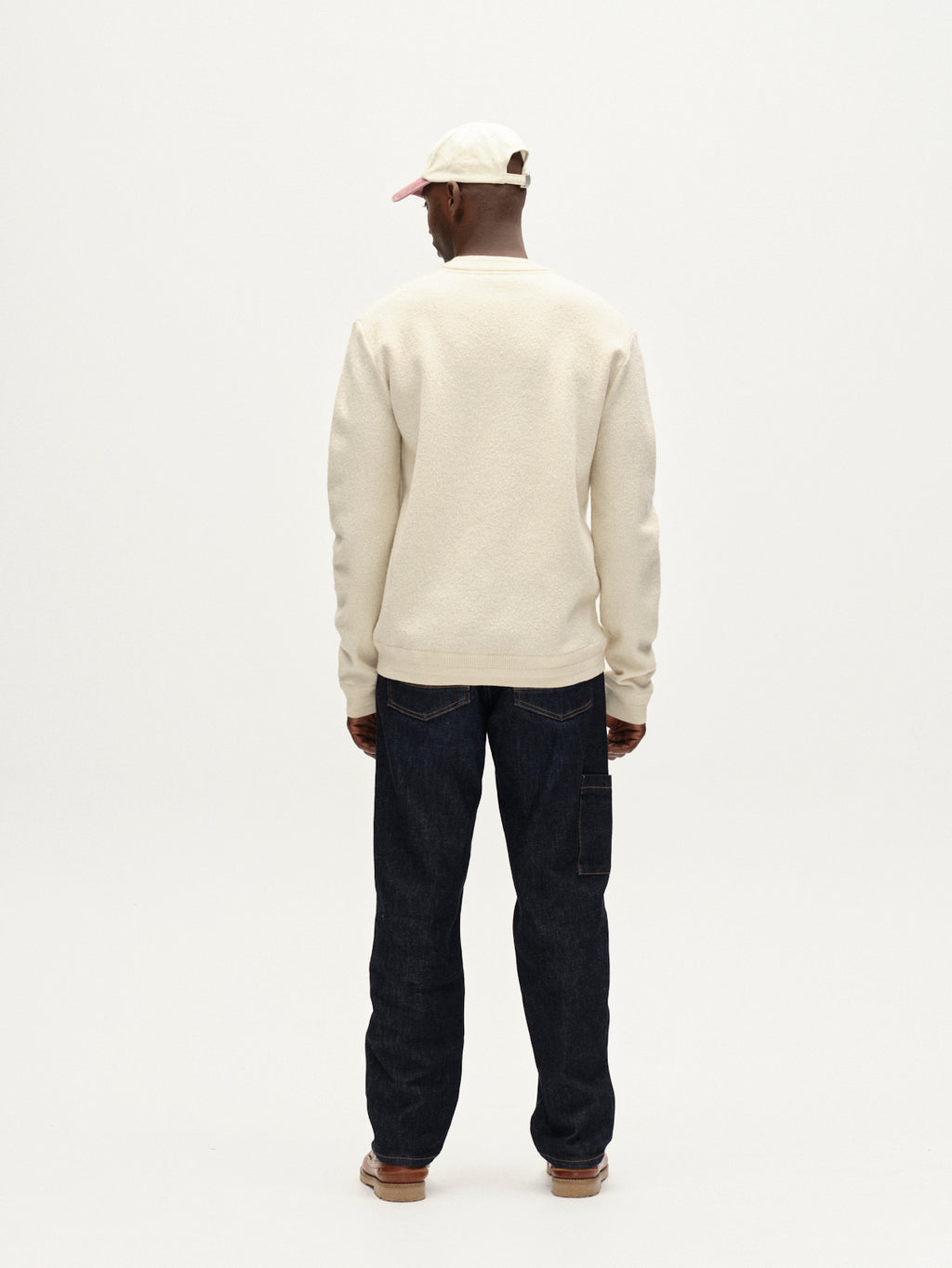 A person stands facing away, wearing the Gabba Bristol Led sweater in White Almond, paired with dark jeans, brown shoes, and a light cap against a plain white background.