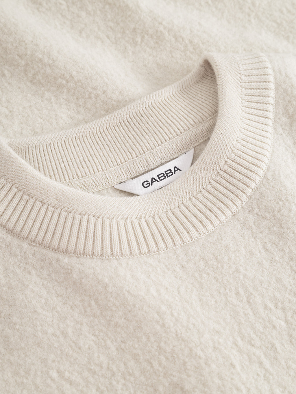 Close-up of the Bristol Led sweater in White Almond by Gabba, featuring a ribbed crew neckline, a white brand label, and soft textured fabric that highlights its cozy wool-blend quality.