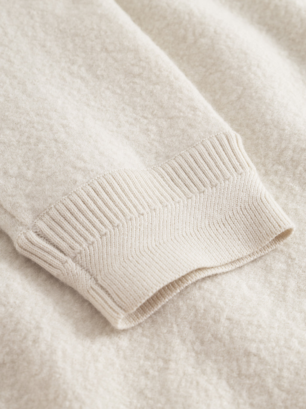 Close-up of the Gabba Bristol Led sweater in White Almond, featuring a ribbed knit cuff and light wool texture, laid flat on a matching surface for a minimalist look.