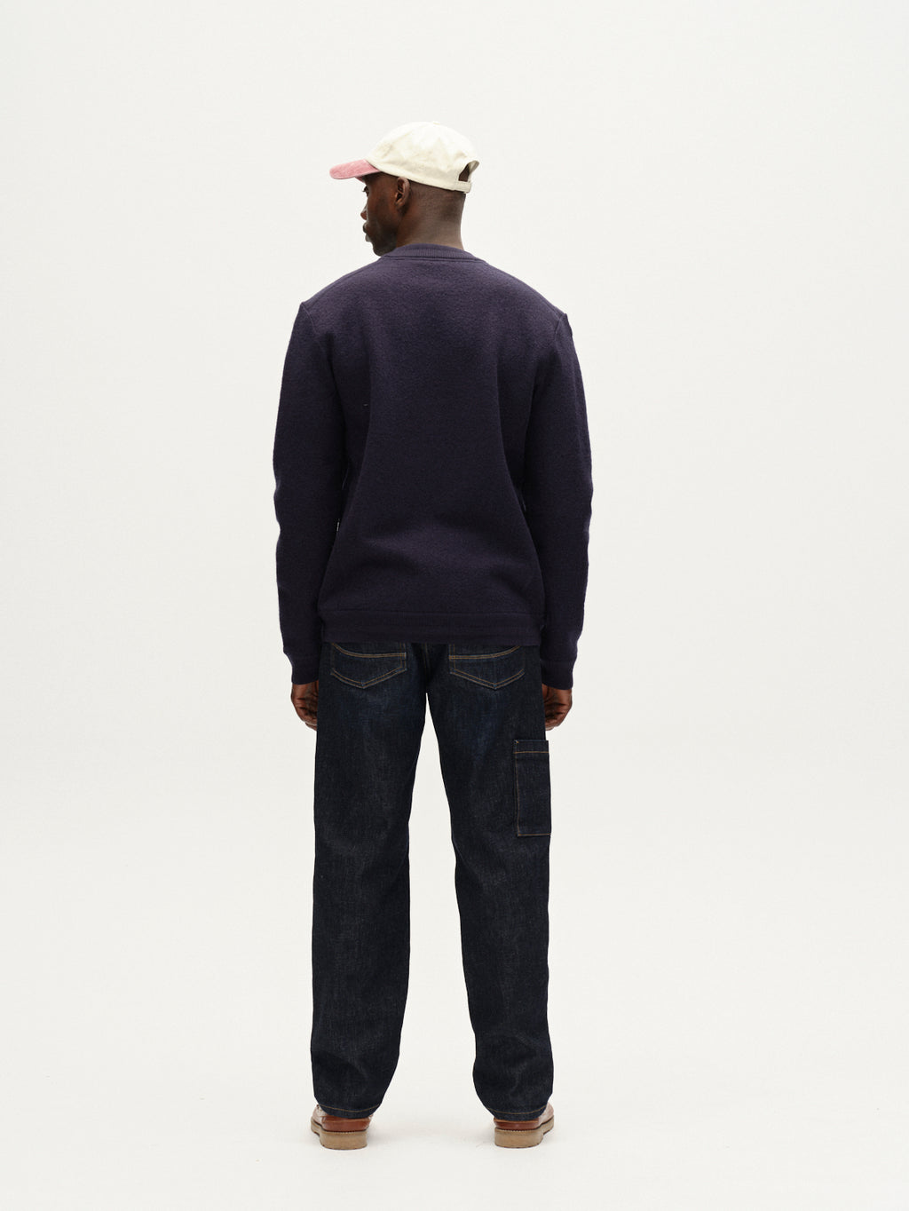 A person stands facing away from the camera, wearing Gabba’s Bristol Led - Navy jeans, a dark purple boiled wool sweater, a cream and pink cap, and light brown shoes against a plain white background.