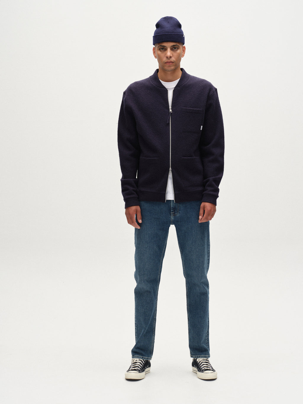 A man stands facing forward against a plain white background, wearing the Gabba Cardiff Led - Navy jacket with a bomber collar and 2-way zip, styled over a white t-shirt, blue jeans, navy beanie, and black-and-white sneakers.
