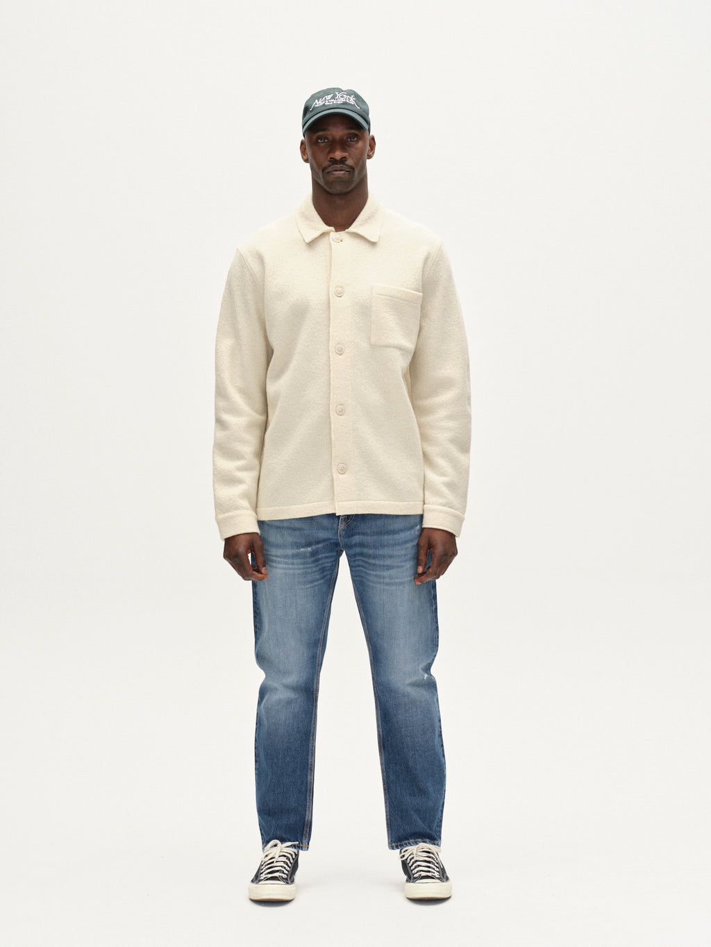 A man stands facing forward against a white background, wearing Gabba's Preston Led - White Almond wool overshirt, blue jeans, black-and-white sneakers, and a green baseball cap.