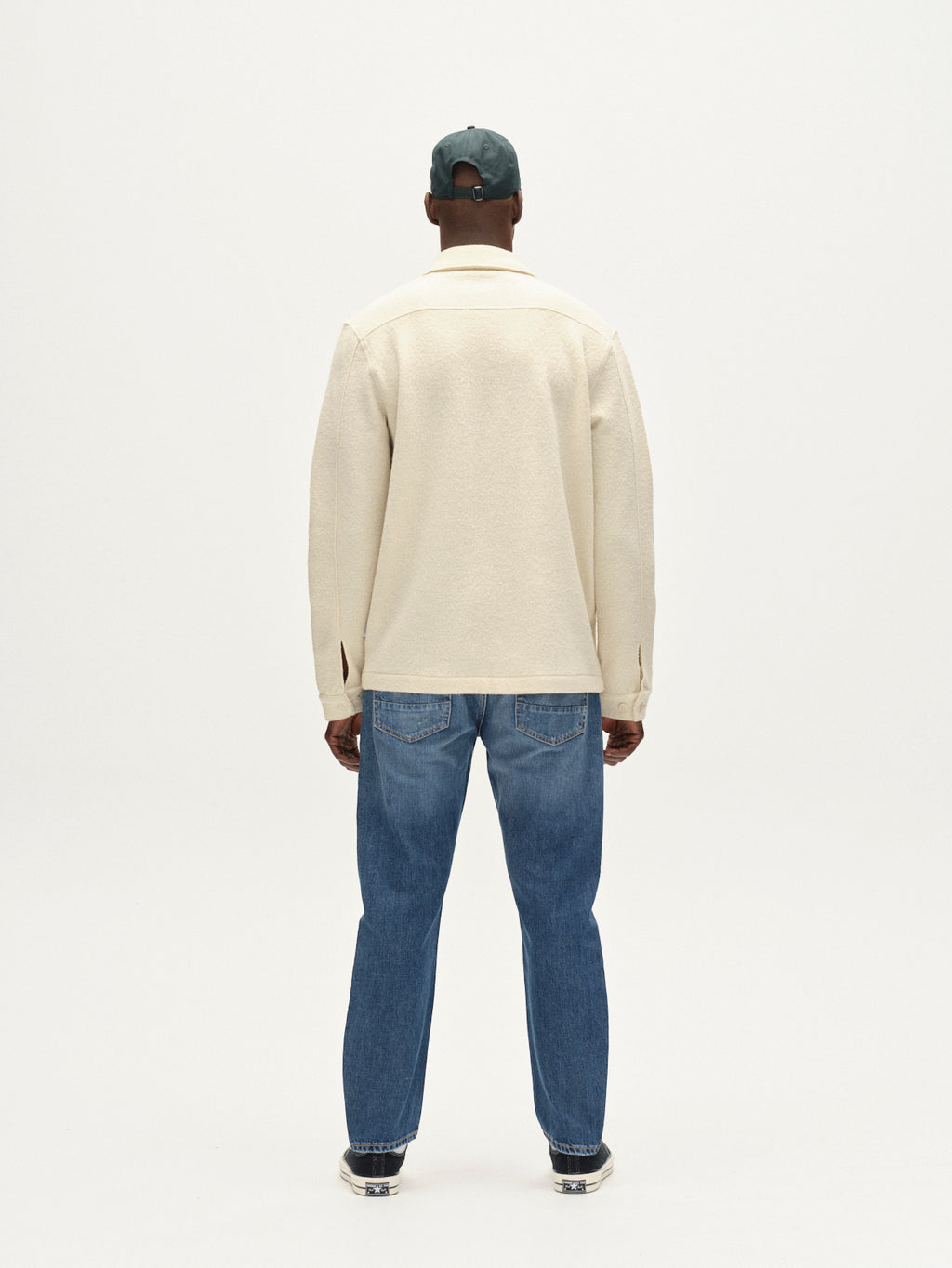 A person faces away in Gabba’s Preston Led - White Almond boiled wool jacket, blue jeans, sneakers, and a green cap against a plain white background.