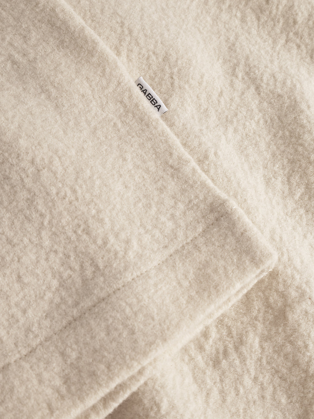 A close-up of the plush, white almond Preston Led blanket by Gabba shows its fluffy texture and a small white label with the brand name sewn onto the tailored edge.