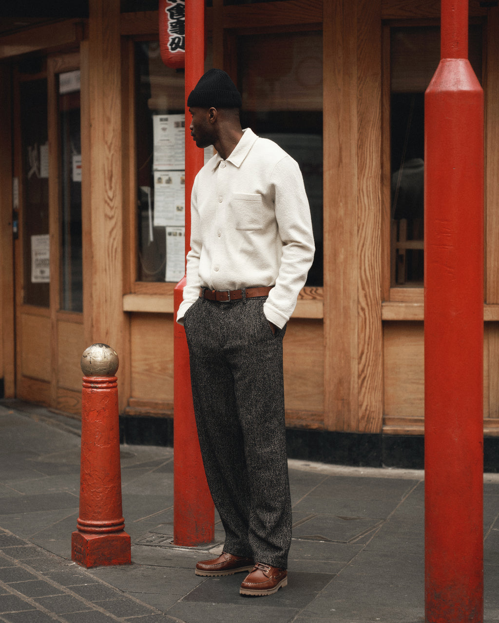 A man wears the Gabba Preston Led - White Almond overshirt, gray textured pants, brown shoes, and a black beanie, standing hands-in-pockets before a wooden building with red pillars, looking to the side.