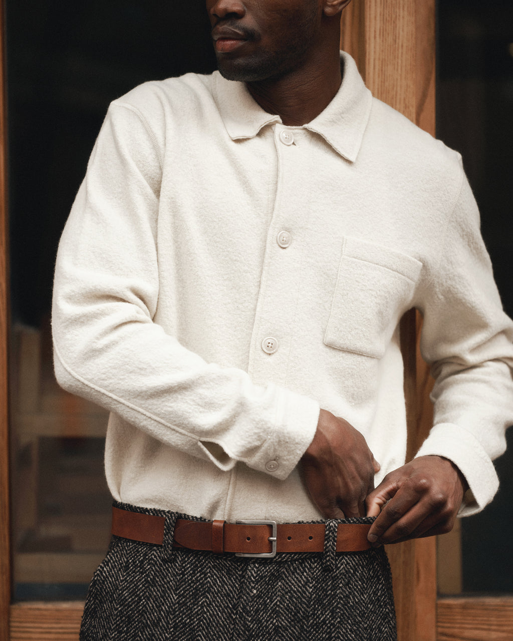 A man adjusts his brown leather belt while wearing the Gabba Preston Led - White Almond shirt and patterned trousers, standing against a backdrop of wooden panels and glass.