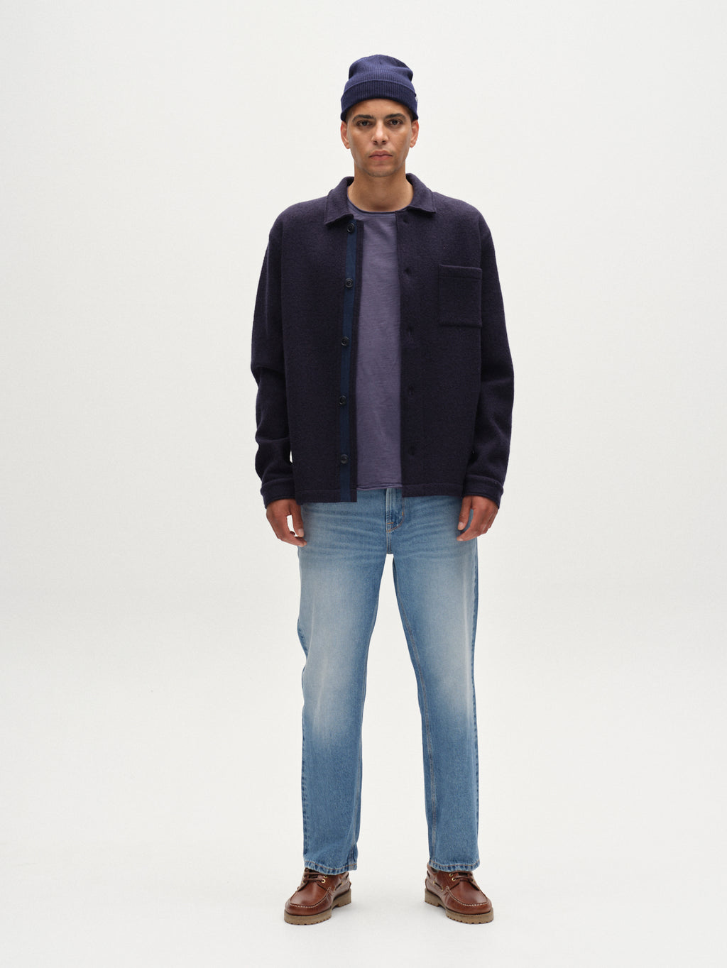A man faces forward against a white background, wearing the Gabba Preston Led - Navy boiled wool overshirt with tailored edges over a purple tee, paired with light blue jeans, a navy beanie, and brown leather shoes.