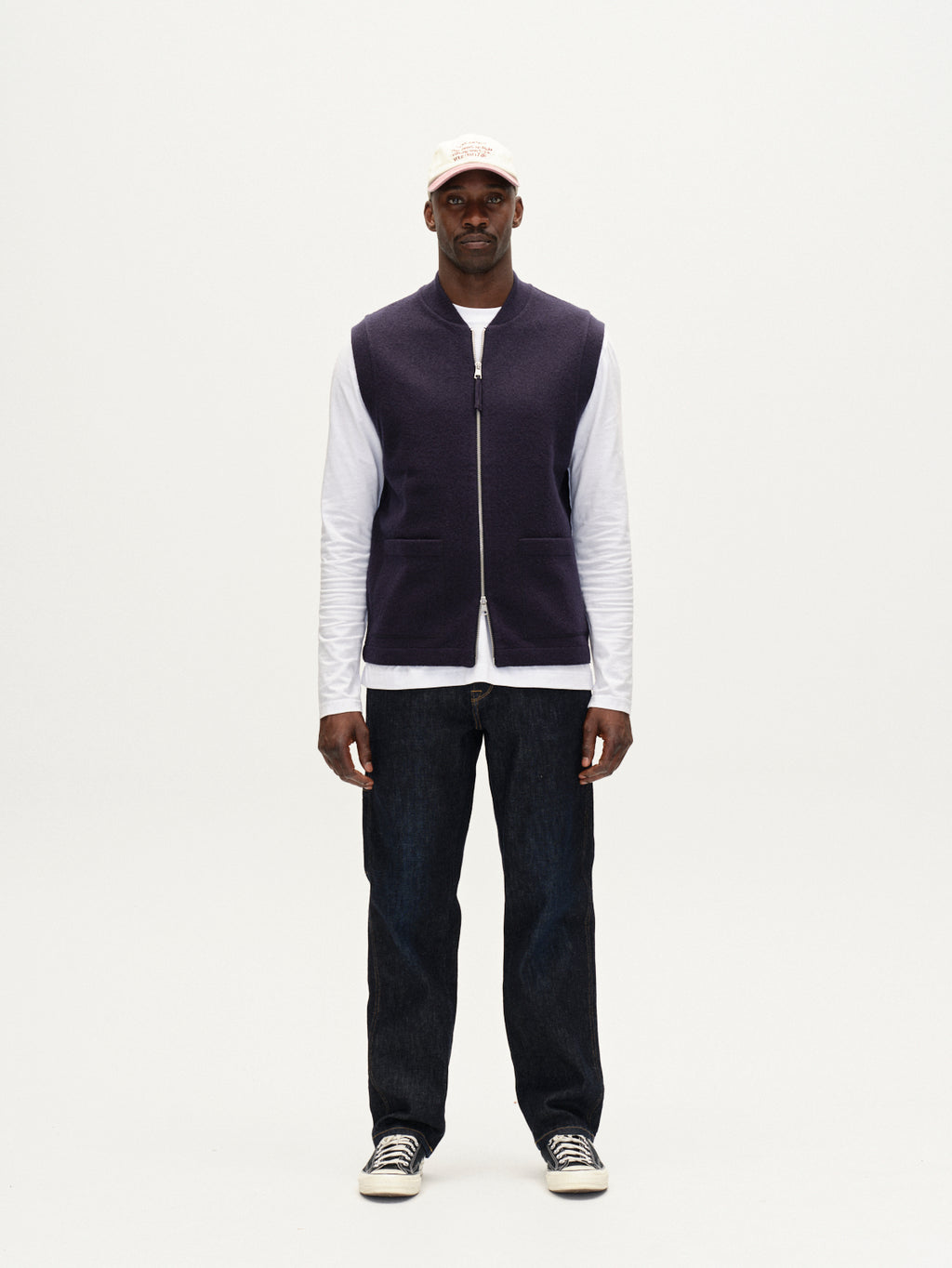 A man stands facing forward against a plain white background wearing the Gabba Norwich Led navy utility vest over a white long-sleeve shirt, paired with a light pink cap, dark jeans, and black sneakers.