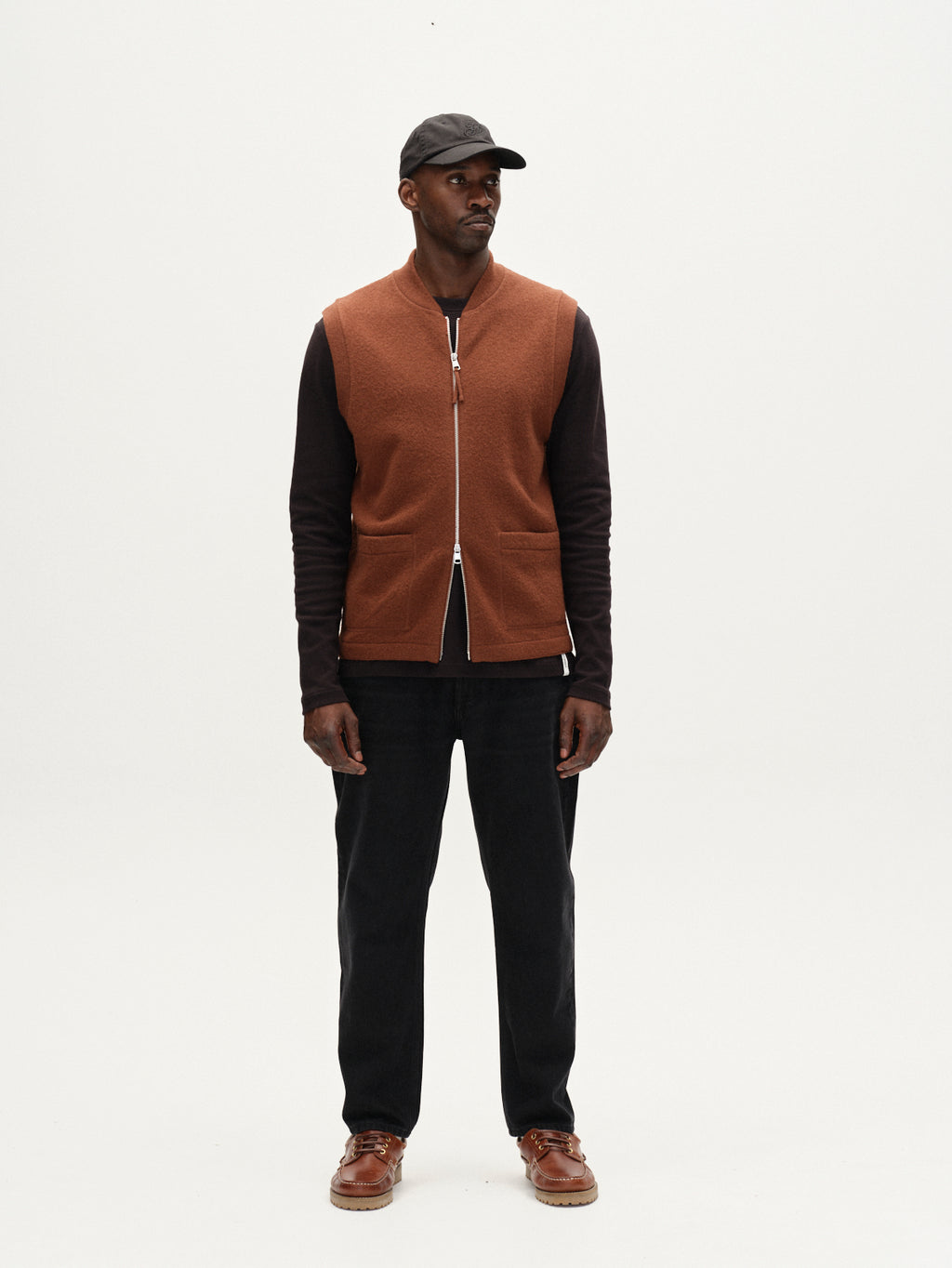 A man stands against a plain white background, wearing the Gabba Norwich Led - Brownish utility vest as a stylish layer over a black long-sleeve shirt, dark pants, and brown shoes. He looks slightly left with a neutral expression, accessorized with a black cap.