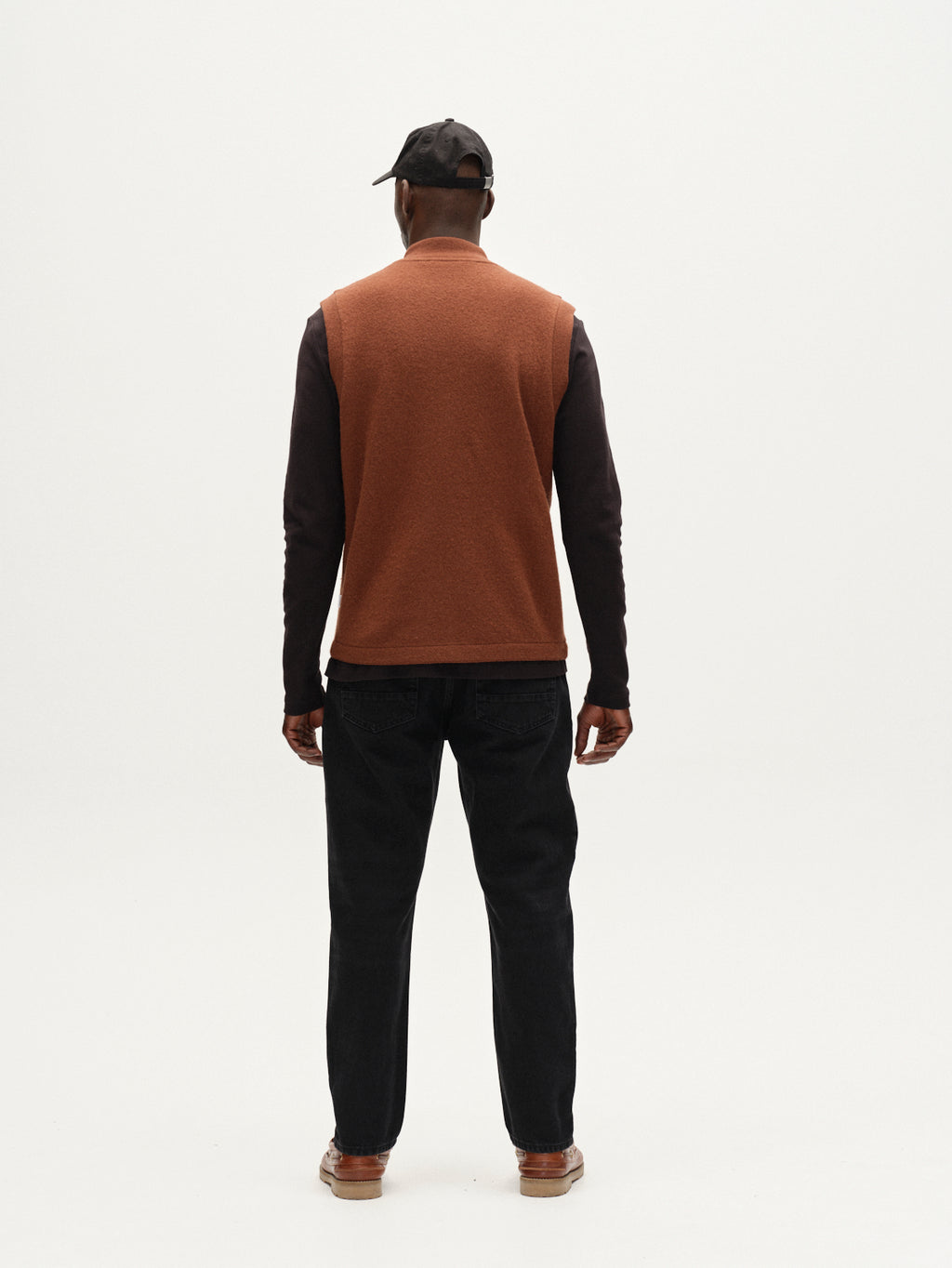 A man faces away from the camera, wearing Gabba's Norwich Led - Brownish utility vest over a long-sleeve shirt, paired with a black cap, dark jeans, and brown shoes against a plain white background.