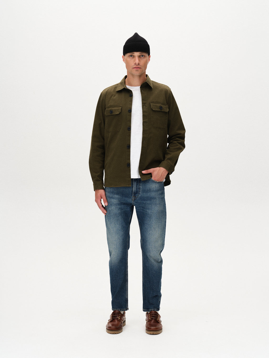 A man stands facing forward, wearing a black beanie, an olive green button-up jacket over a white t-shirt, dark blue Carl Oskin denim jeans by Gabba, and brown shoes against a plain white background.