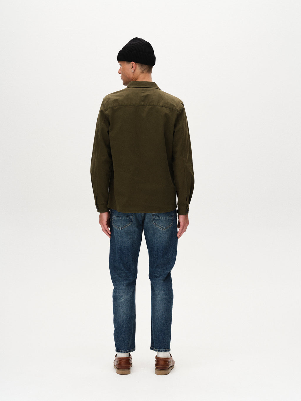 A man stands facing away from the camera in a relaxed fit olive green jacket, Carl Oskin - Dark Blue Denim jeans by Gabba, brown shoes, and a black beanie, showcasing effortless casual style against a plain white background.