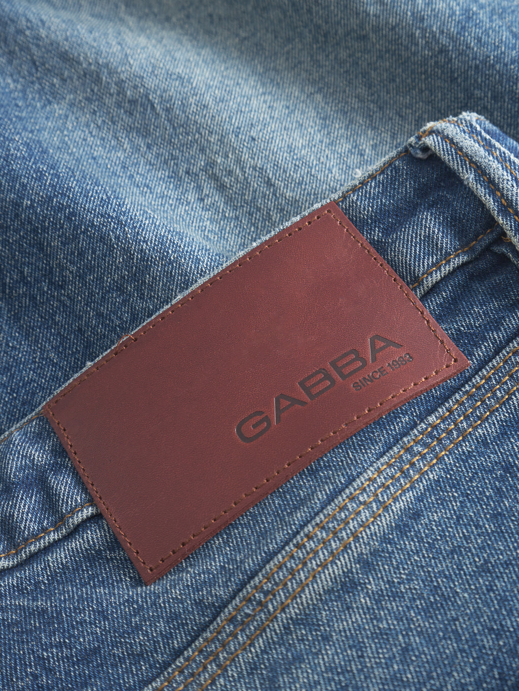Close-up of Carl Oskin dark blue denim jeans by Gabba with a brown leather patch embossed GABBA Since 1983. The contrasting dark stitching on faded fabric embodies a casual style complementing their loose tapered fit.