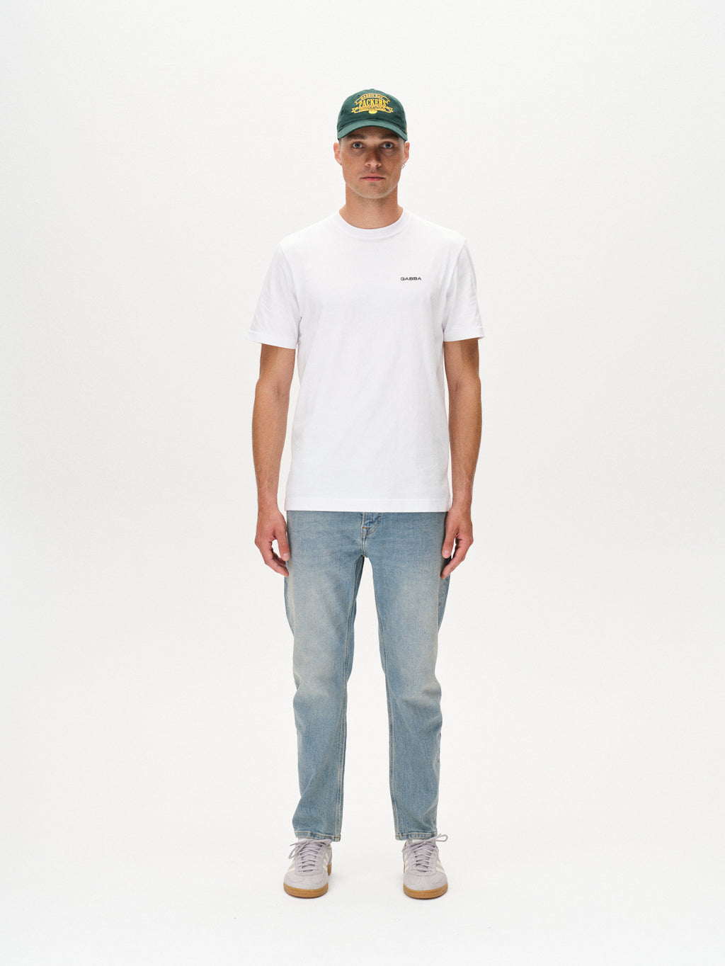 A person stands facing forward in a green cap, white tee, and Gabbas Carl Cros Lt. Blue Denim jeans with white sneakers against a plain background, highlighting a casual contemporary style.