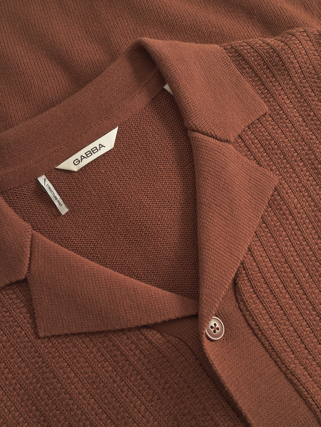 A close-up of the Sakai Wong LS cardigan by Gabba in brown rust, showcasing a resort-style collar with button, signature neckline tag, and fabric with ribbed texture on one side and smooth knit on the other.