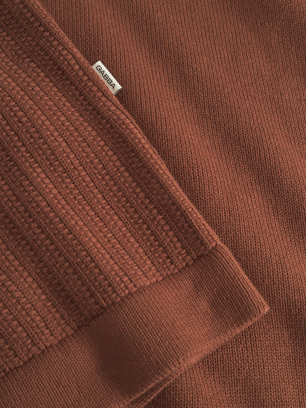 Close-up of the Sakai Wong LS cardigan by Gabba in brown rust, showcasing ribbed and smooth cotton knit textures. A small white tag with Gabba is sewn into the seam.