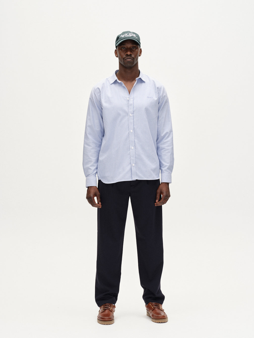 A man stands facing forward in a Gabba Sven Reg Crest LS - Blue Stripe shirt, dark trousers, brown shoes, and a dark baseball cap against a plain white background.