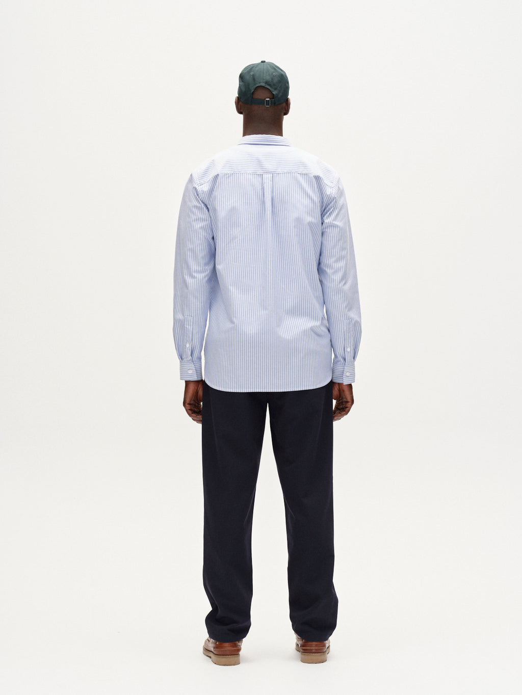 A person stands with their back to the camera wearing Gabbas Sven Reg Crest LS - Blue Stripe shirt, a green baseball cap, dark pants, and brown shoes against a plain white background.