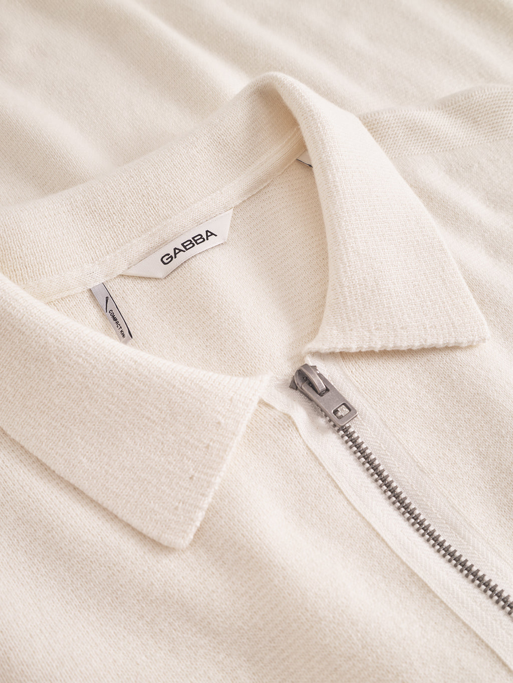 Close-up of the Gabba Blanko New Zip LS in off white, a cream-colored half-zip knit shirt made from a textured cotton-wool blend, featuring a silver zipper and a visible Gabba label stitched inside the collar.