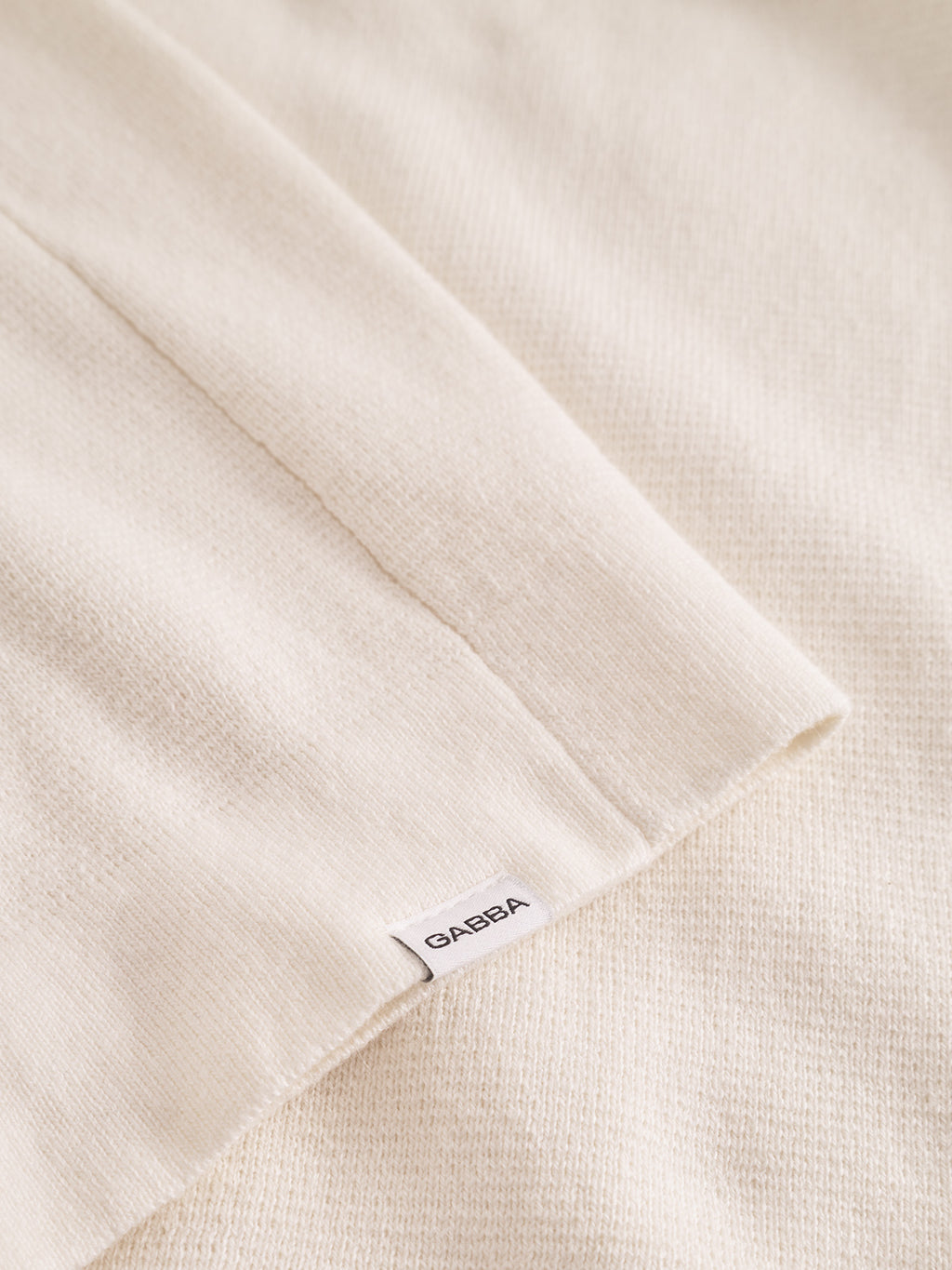 A close-up of the Blanko New Zip LS in off white, a soft cotton-wool blend knit from Gabba, shows a neatly finished edge and a small white tag with