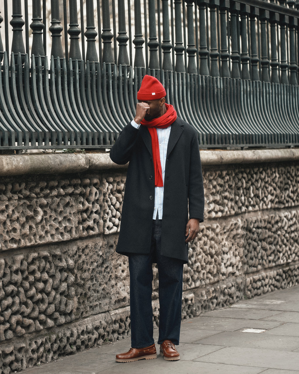 A man stands on a city sidewalk by a stone wall and black fence, wearing a dark coat, jeans, brown shoes, red scarf, and the Gabba Biella Aico - Red ribbed knit beanie while adjusting his glasses.