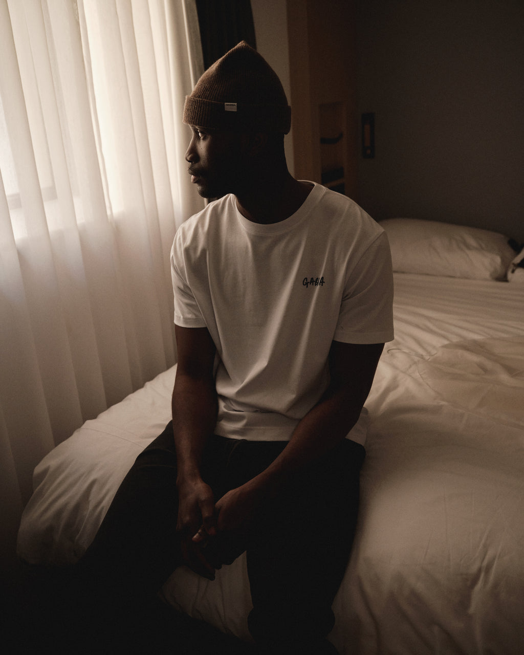 A man wearing a white t-shirt and the Gabba Biella Aico - Dk. Brown beanie sits on a bed, gazing out a window with sheer curtains in a softly lit, neutral-toned room.