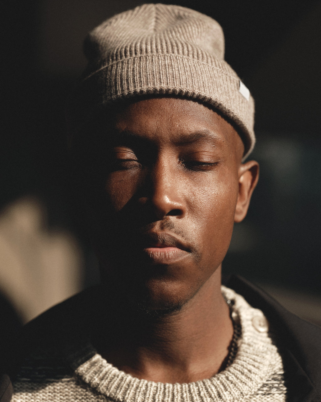 A man wearing the Gabba Biella Aico - Lt. Brown merino wool beanie and a textured sweater stands with his eyes closed, softly lit as shadows partially cover his face.