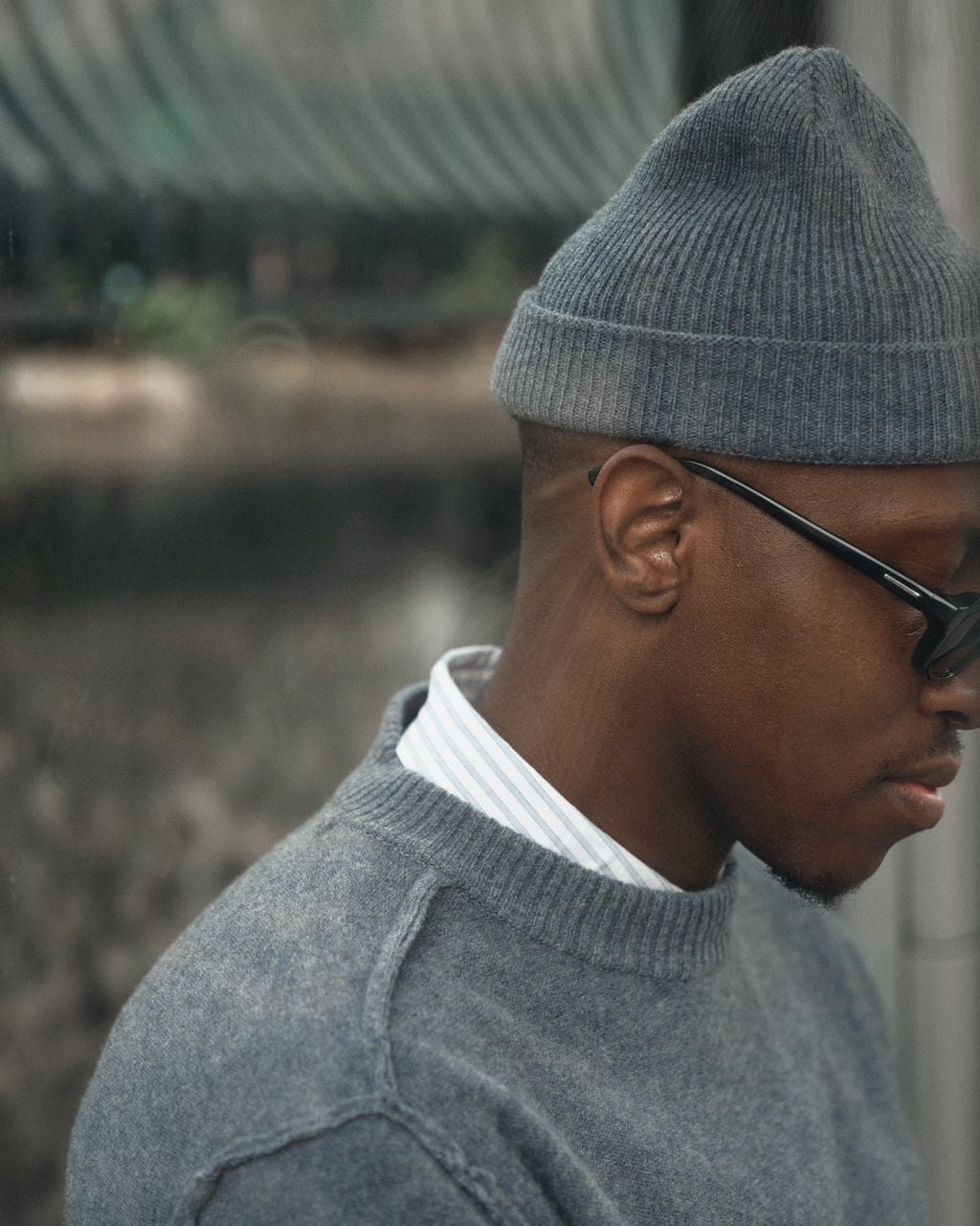 A man wearing the Gabba Biella Aico Vintage Indigo ribbed beanie, a gray sweater, striped collared shirt, and dark sunglasses stands outdoors, looking down with a blurred background behind him.