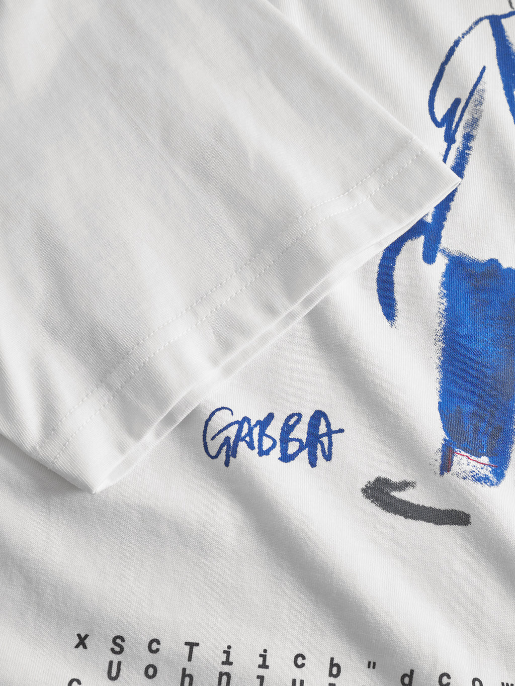 A close-up of the Gabba Dune Kay Man SS GOTS - White tee, featuring blue abstract artwork, GABBA handwritten, and partial black text along the bottom edge. The sleeve appears in the upper left. Part of the GOTS-certified collection.