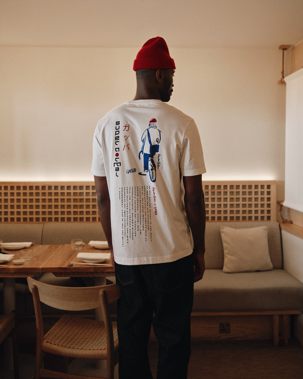 A man wearing a red beanie and the Gabba Dune Kay Man SS GOTS tee in white stands in a cozy, modern dining area with wooden furniture and neutral tones, facing away from the camera.