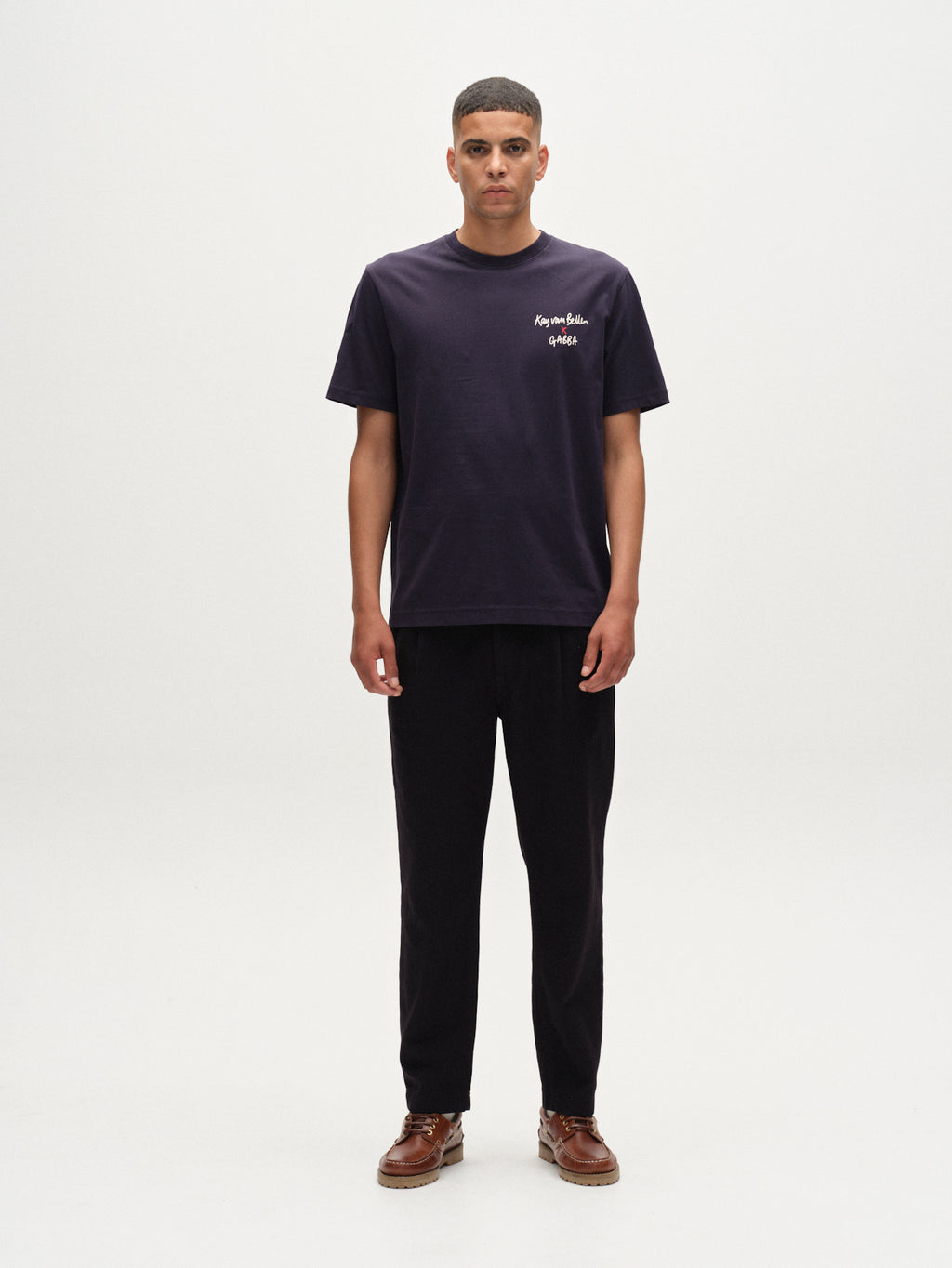A man faces forward against a plain white background, wearing Gabba’s Dune Kay Man SS GOTS tee in navy with small white text on the upper left chest, paired with dark pants and brown shoes.