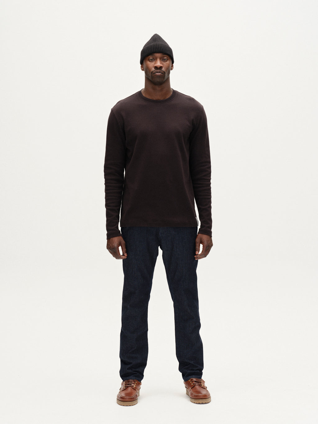 A man stands facing forward against a plain white background, wearing a black beanie, a dark long-sleeve shirt, brown shoes, and Gabbas Paul Sisu jeans in dark denim.