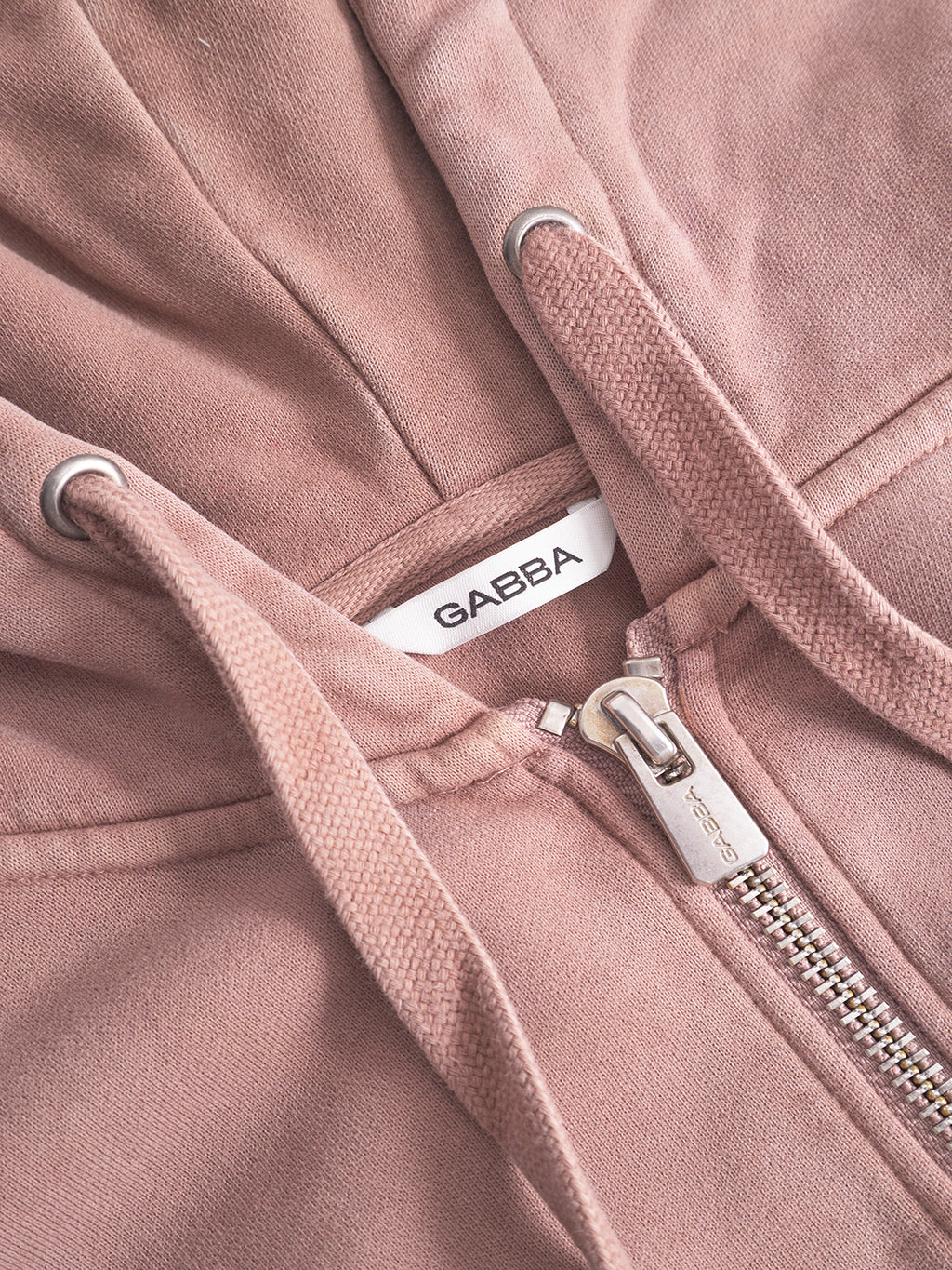 Close-up of the Delphi Bump GWC - Rose hoodie by Gabba, featuring a zipper, metal eyelets, and drawstrings. The Gabba black label is visible inside the collar, enhancing its modern casual appeal.