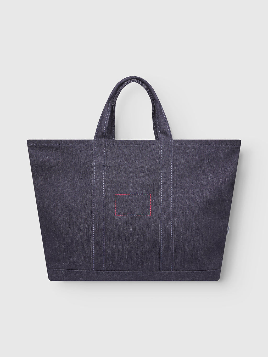 The Gabba Bag Self - Dark Blue Denim is a spacious tote made from dark blue denim, featuring two handles, visible stitching, and a small front rectangular patch with red stitches, shown against a plain light background.