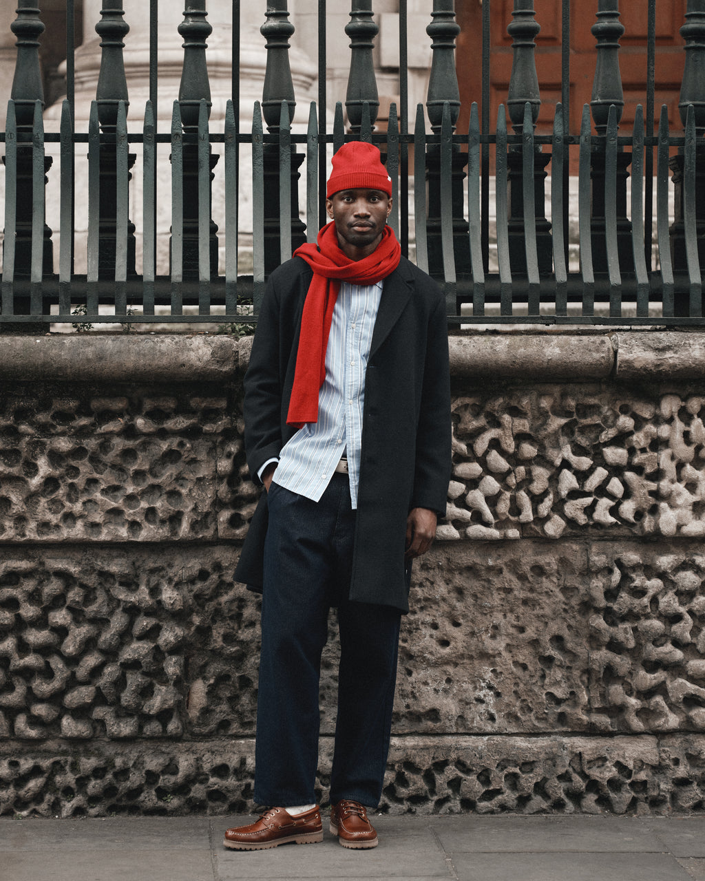 Wearing the Gabba Biwa Aico - Red beanie, a person stands in front of a stone wall and iron fence on a city street, paired with a merino wool scarf, black coat, light blue shirt, dark pants, and brown shoes.