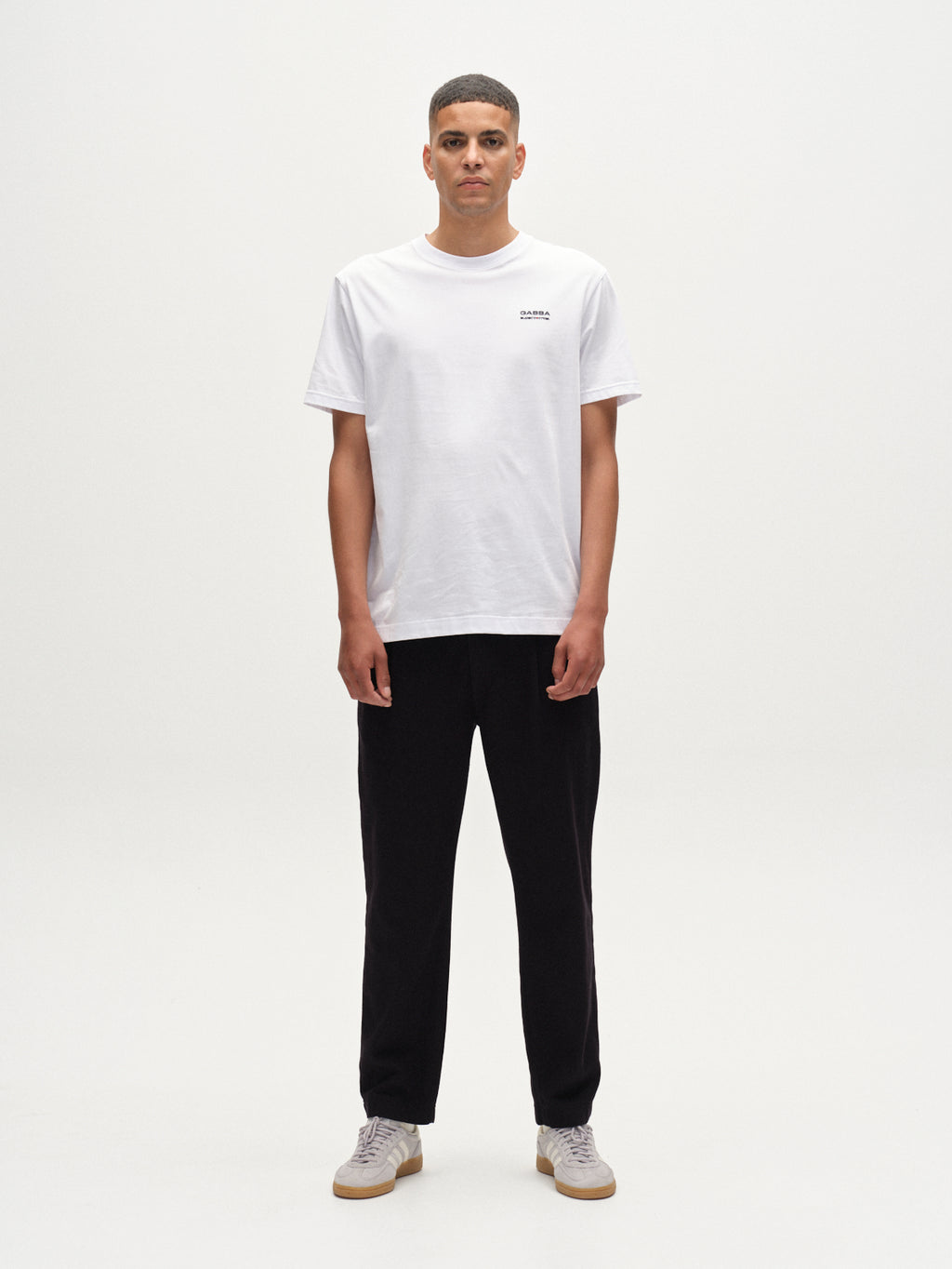 A person faces the camera against a plain white background, wearing Gabba’s Dune Kay Man Bauw SS GOTS T-shirt in white with a small left-chest logo, paired with black pants and light-colored sneakers.