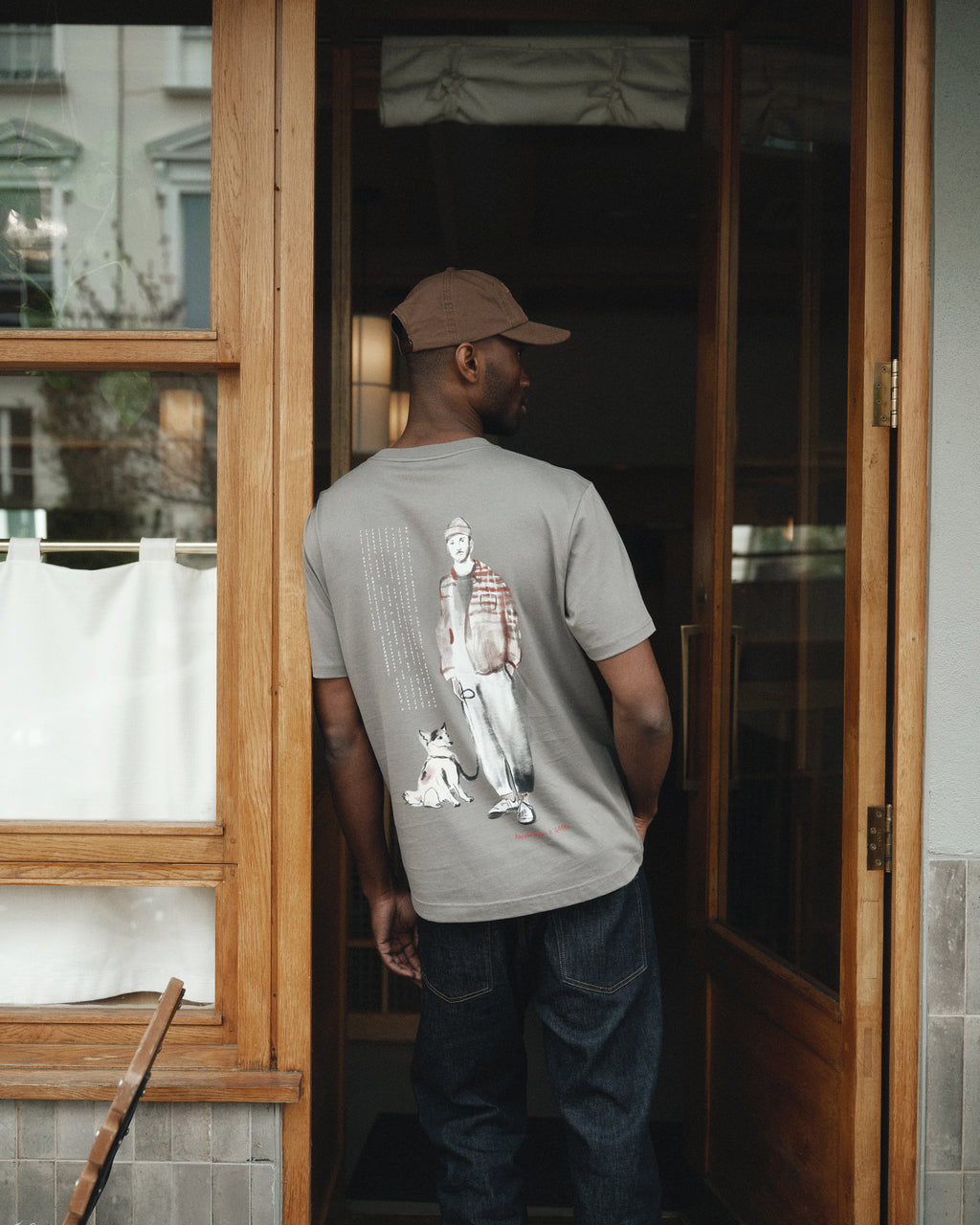A man in a brown cap and a Gabba Dune Kay Man Bauw SS GOTS T-shirt in Dust Sage Green with an illustrated back stands in a doorway. The wood-framed door and windows have partially visible white curtains.