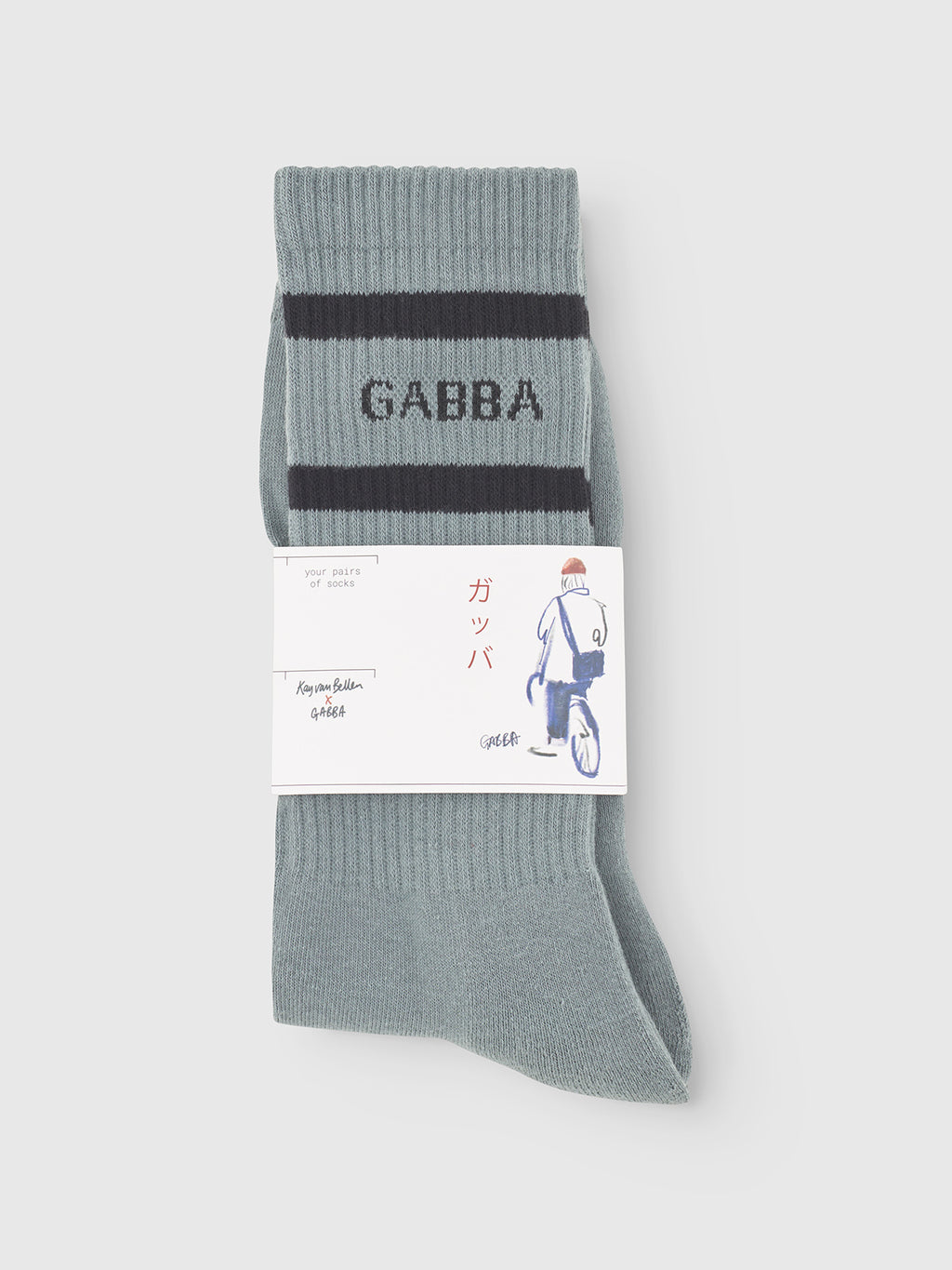 A pair of folded Zino Socks in Dust Sage Green by Gabba, featuring two dark stripes and GABBA text, packaged with a white label with Japanese writing and a bicycle illustration. Made from a soft cotton blend for daily comfort.