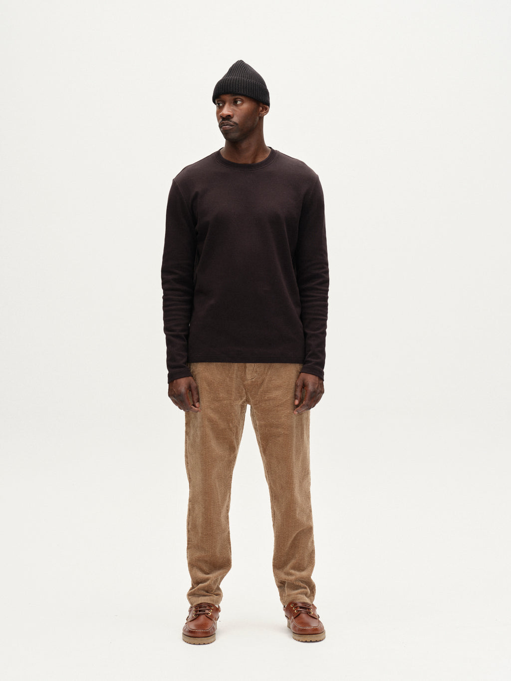 A man stands against a plain white background wearing the Gabba Monza Ibre - Sand corduroy trousers, paired with a black beanie, dark long-sleeve shirt, and brown shoes. He faces forward, hands by his sides, exuding casual sophistication.